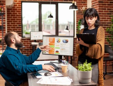Young woman stands in front of colleague, holding clipboard and confidently explaining project updates. Supportive bearded man listening to female coworker, ready to collaborate on company research.
